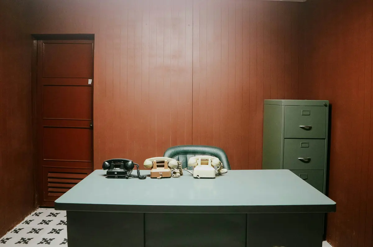 Escritorio de admisión con tres teléfonos antiguos y un archivador metálico verde al lado, en una habitación con paredes de madera.