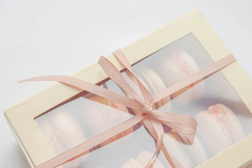 Caja blanca de pastelería con ventana transparente, atada con una cinta rosa, con macarons dentro.