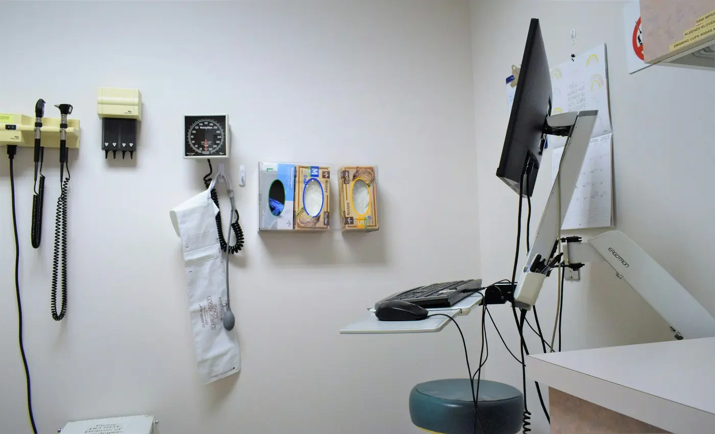 Pared de un consultorio con el otoscopio y el tensiómetro colgados en su soporte, un reloj, cajas de guantes y una pantalla sobre la mesa de trabajo.