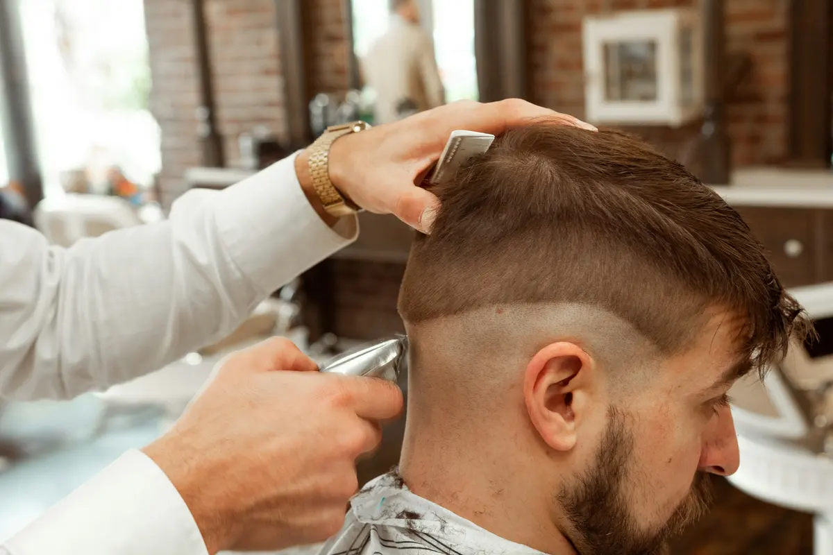 Las manos de un barbero perfilando la nuca de un cliente con la máquina, con luz de día y una pared de ladrillo detrás.
