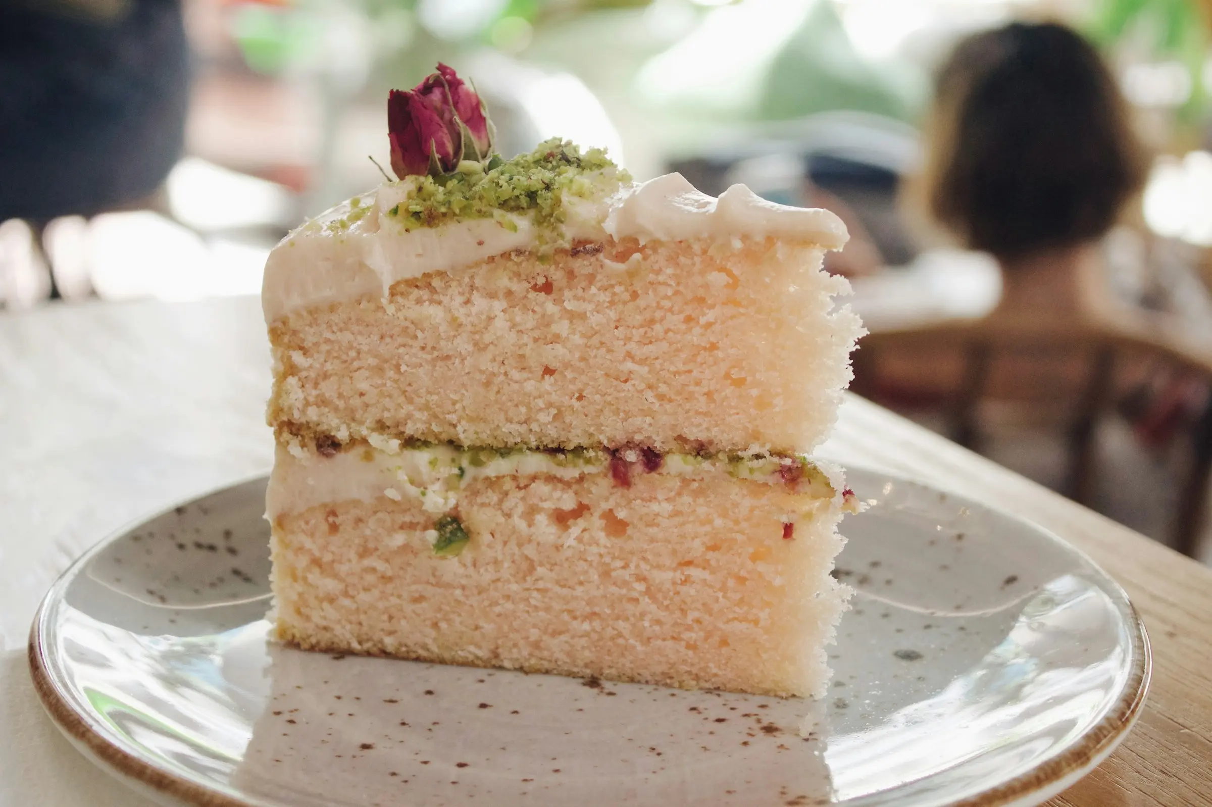 Porción de torta de dos pisos de bizcocho rosado con relleno de pistacho, coronada con crema, pistacho picado y un botón de rosa, en un plato de vidrio.