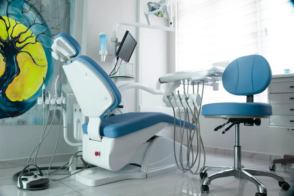 Sillón dental azul con su brazo de instrumentos y el taburete del odontólogo, junto a un cuadro pintado en la pared.
