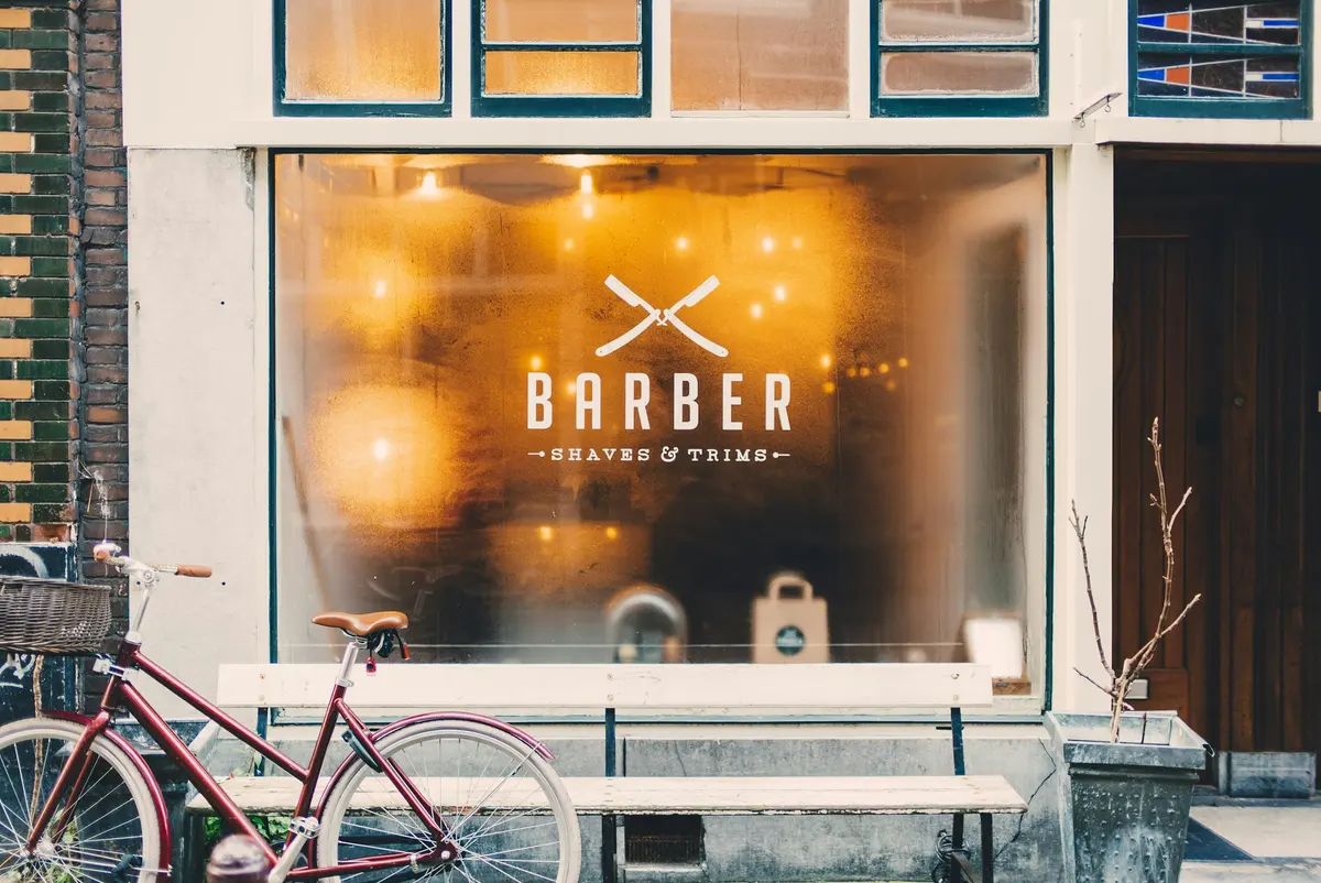 Fachada de una barbería a pie de calle con el rótulo pintado en el escaparate y una bicicleta aparcada delante.
