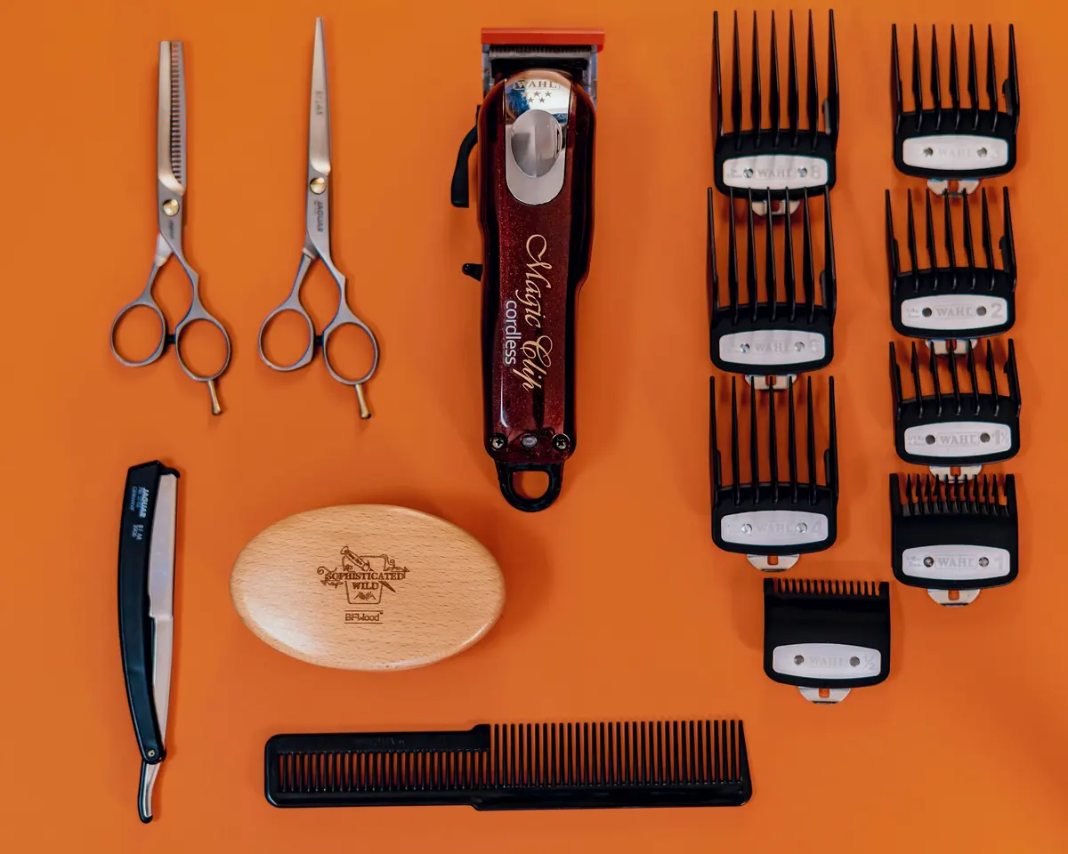 Herramientas de barbería ordenadas sobre una superficie naranja: una máquina de cortar, la fila de peines de medida, dos tijeras, una navaja, un peine y una brocha de afeitar.