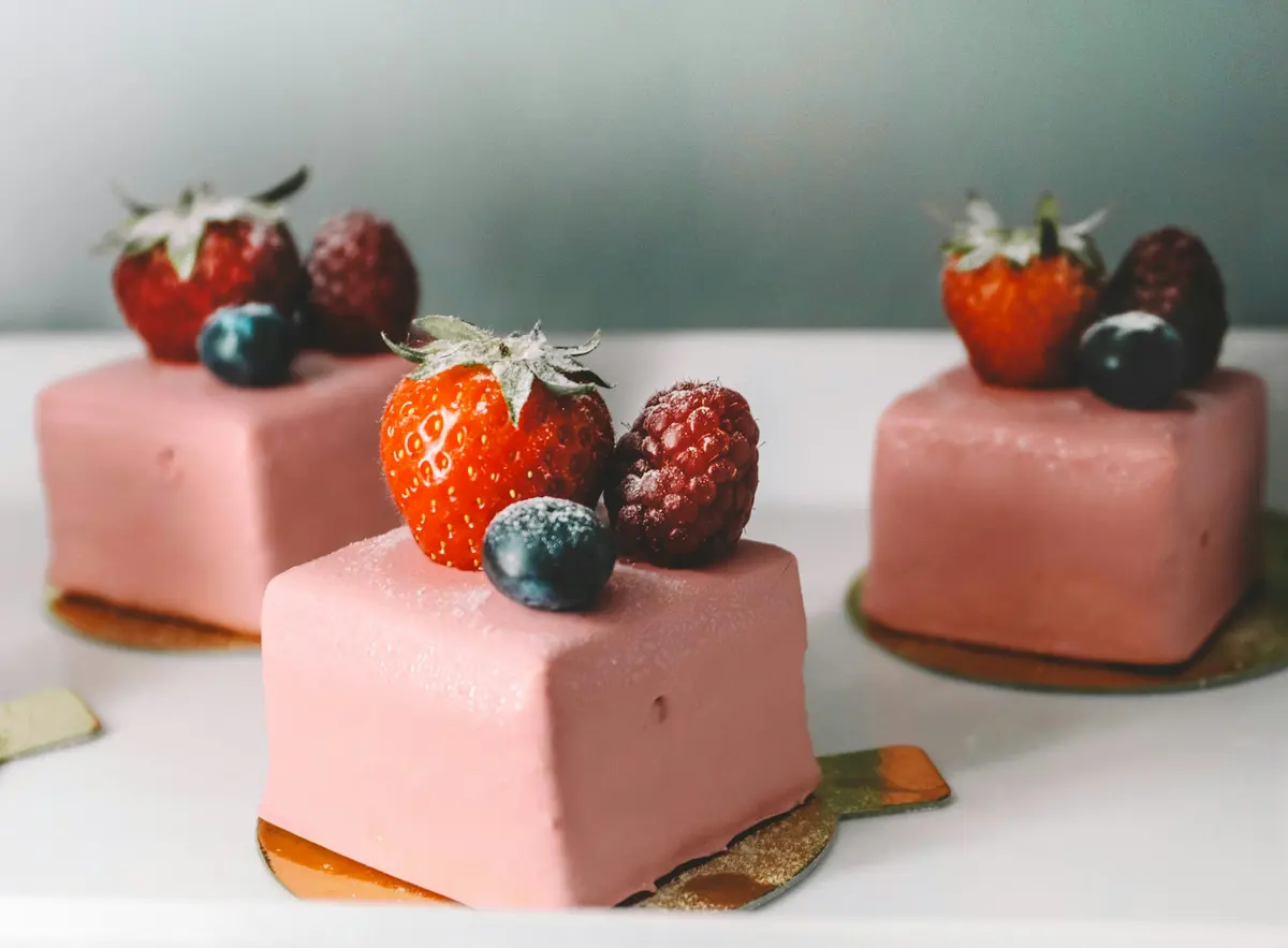 Tres pasteles individuales cuadrados con glaseado rosa, coronados con fresas, frambuesas y arándanos.