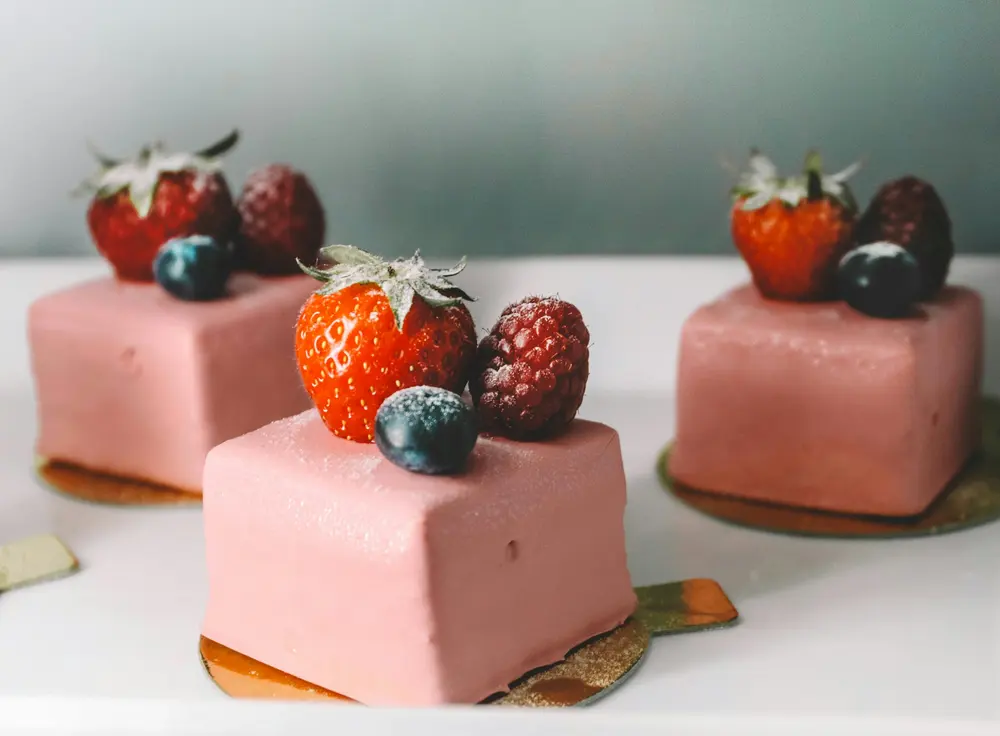 Tres pasteles individuales cuadrados con glaseado rosa, coronados con fresas, frambuesas y arándanos.