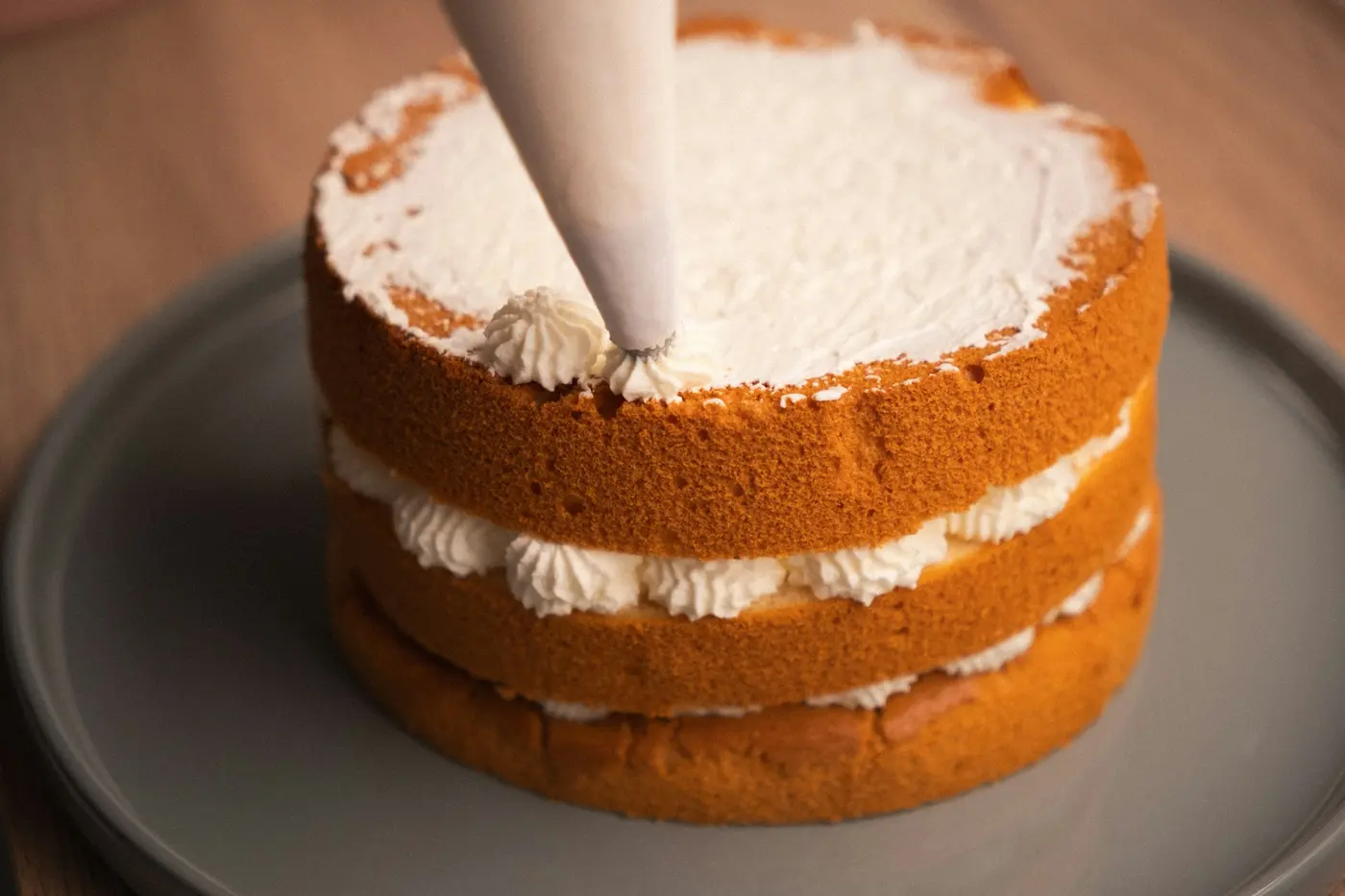 Una manga pastelera aplica crema sobre un bizcocho de dos pisos que muestra los rosetones de relleno entre las capas.
