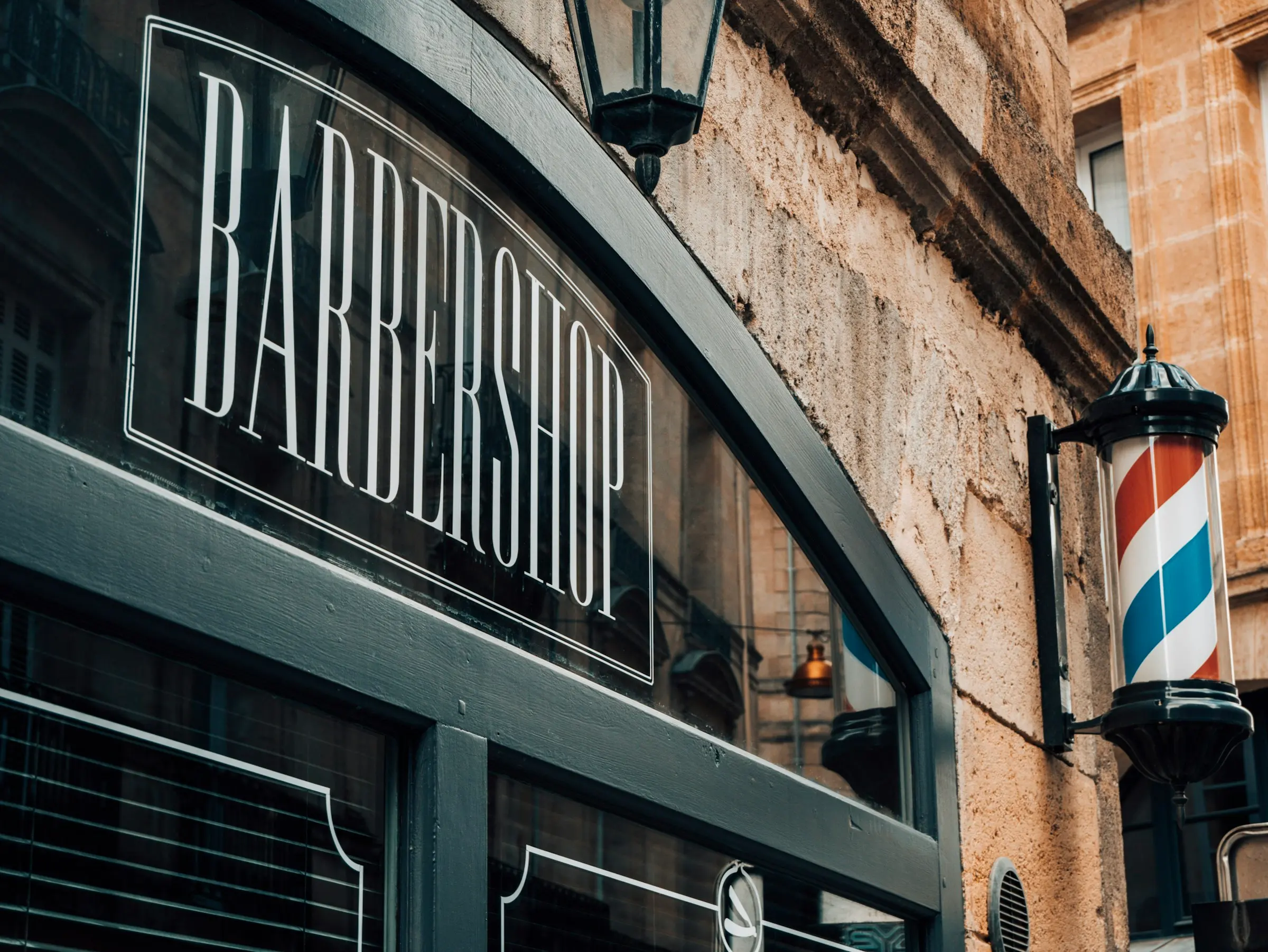 La palabra BARBERSHOP pintada a mano en letras blancas sobre una pared de ladrillo, con un poste de barbero de rayas montado al lado.